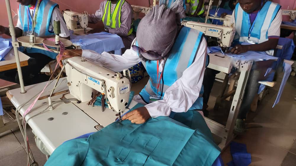 Trainee in headwrap carefully sewing blue fabric on industrial machine